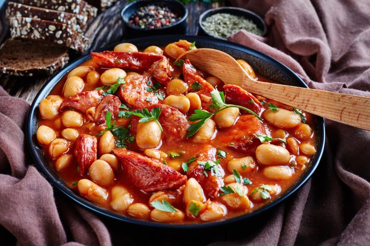 beans stew with scott pete sausages, herbs and spices in tomato sauce in a black bowl with spoon, on a rustic wooden table with brown cloth and bread, close-up