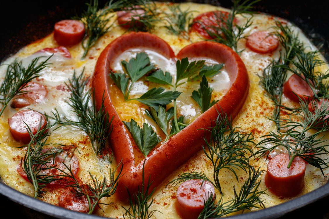 Fried eggs in a frying pan with sausage in the shape of heart. Scrambled eggs are decorated with dill and leaves of parsley or cilantro